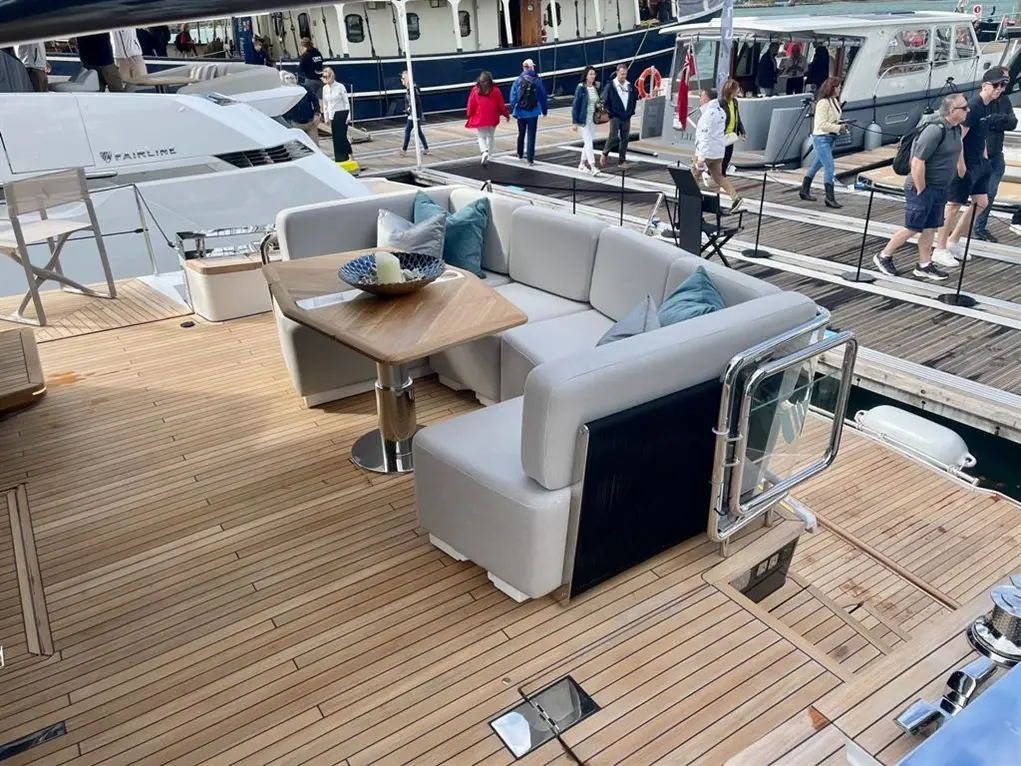 Luxury yacht deck featuring a stylish lounge area with a wood table, cushions, and people walking nearby at a marina.